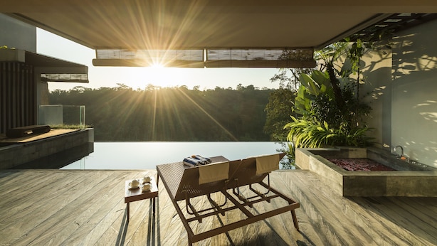 A modern outdoor deck with a wooden table and lounge chairs, overlooking a body of water and a lush garden. - Ayuterra Resort (Ubud)