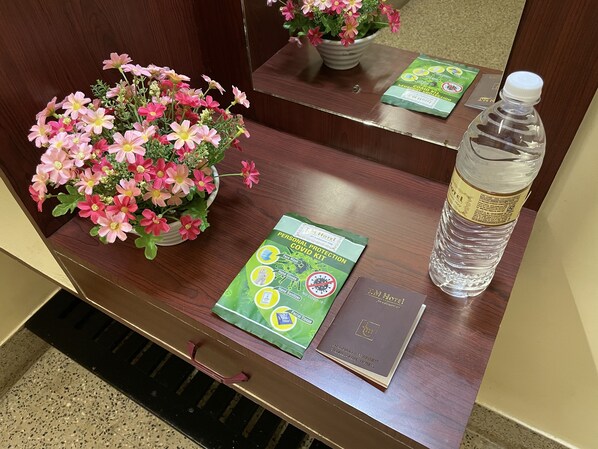 A hotel room desk with a flower arrangement, a personal protection COVID kit, a water bottle, and a welcome booklet.
