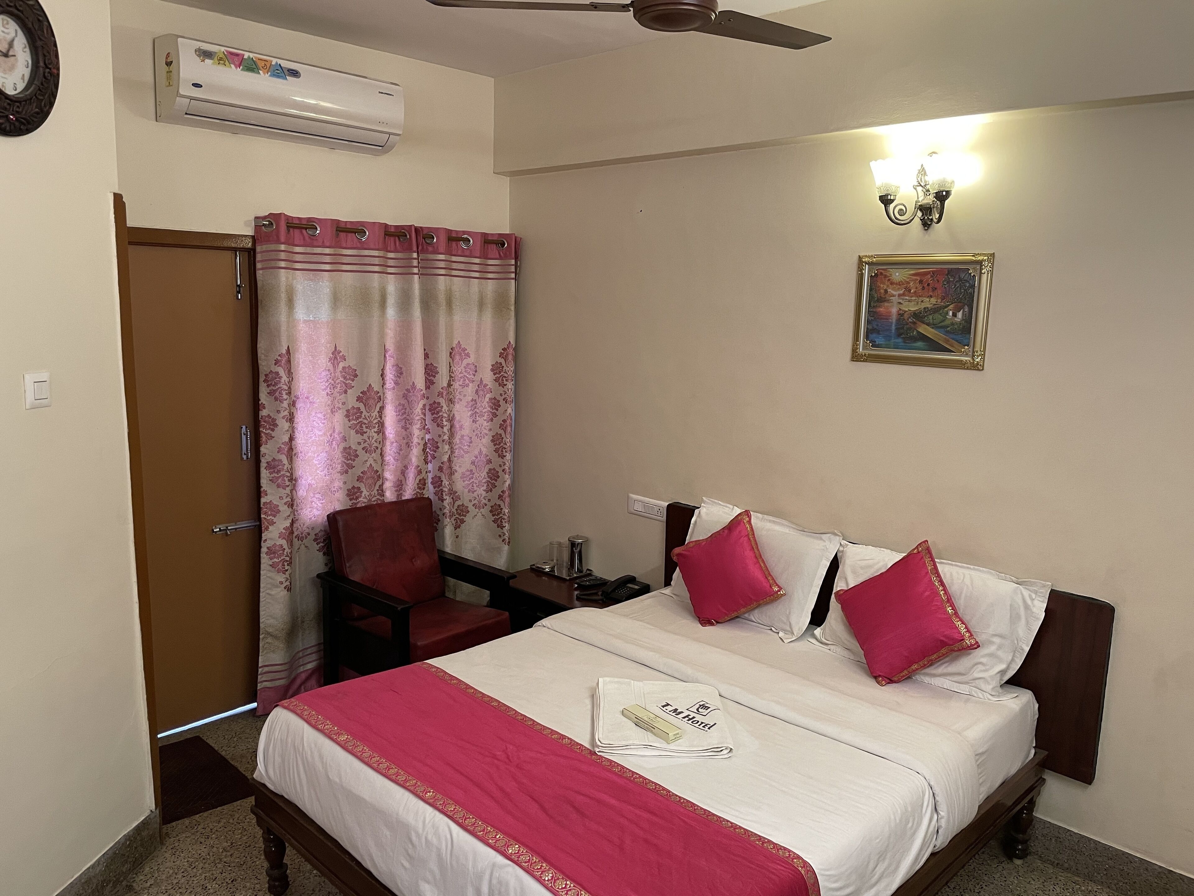 A hotel room with a bed, a chair, a desk, an air conditioner, a ceiling fan, a wall-mounted light, and a painting.