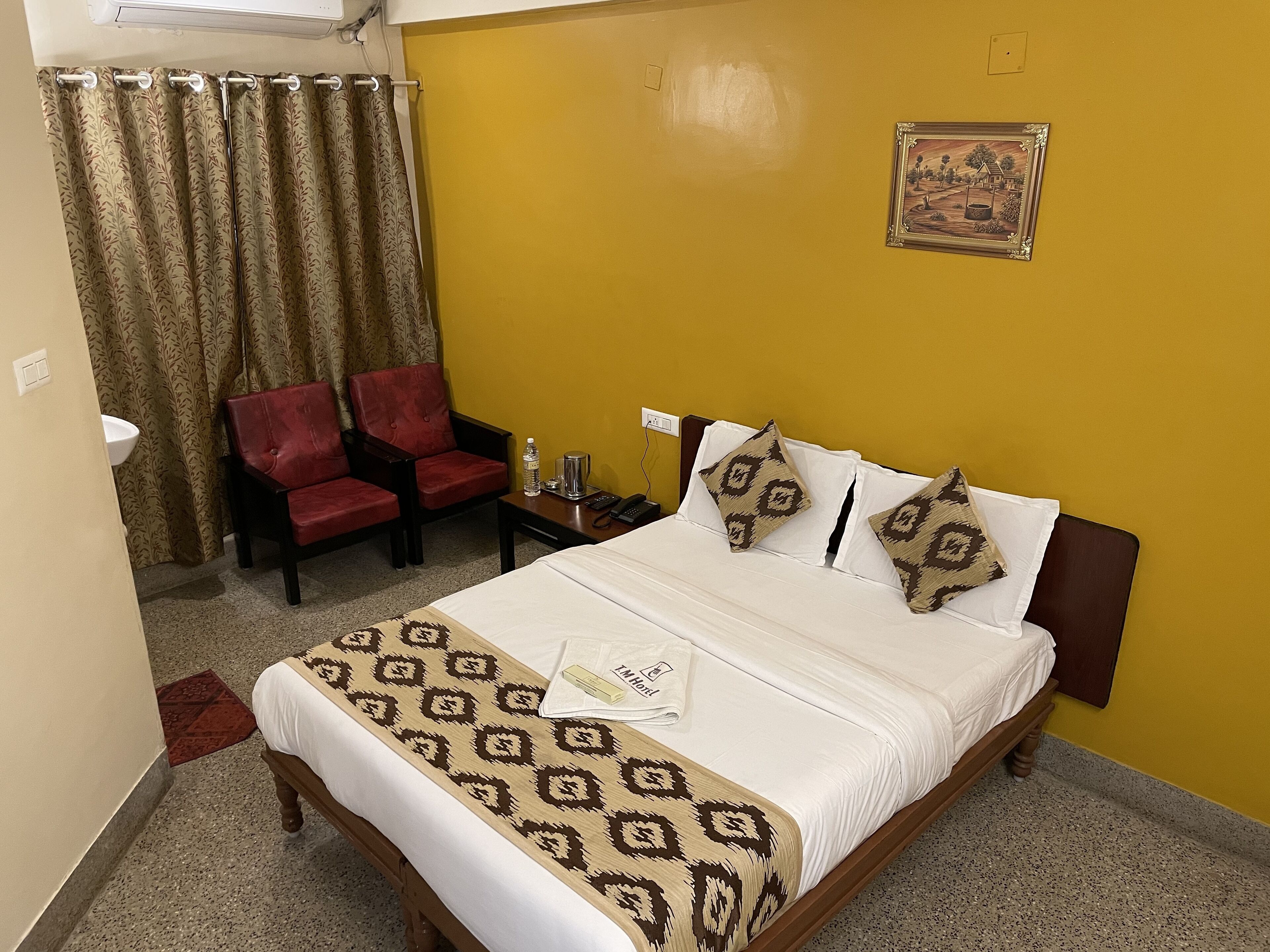 A hotel room with a bed, two red chairs, a small table with a water bottle and a coffee pot, and a picture on the wall.