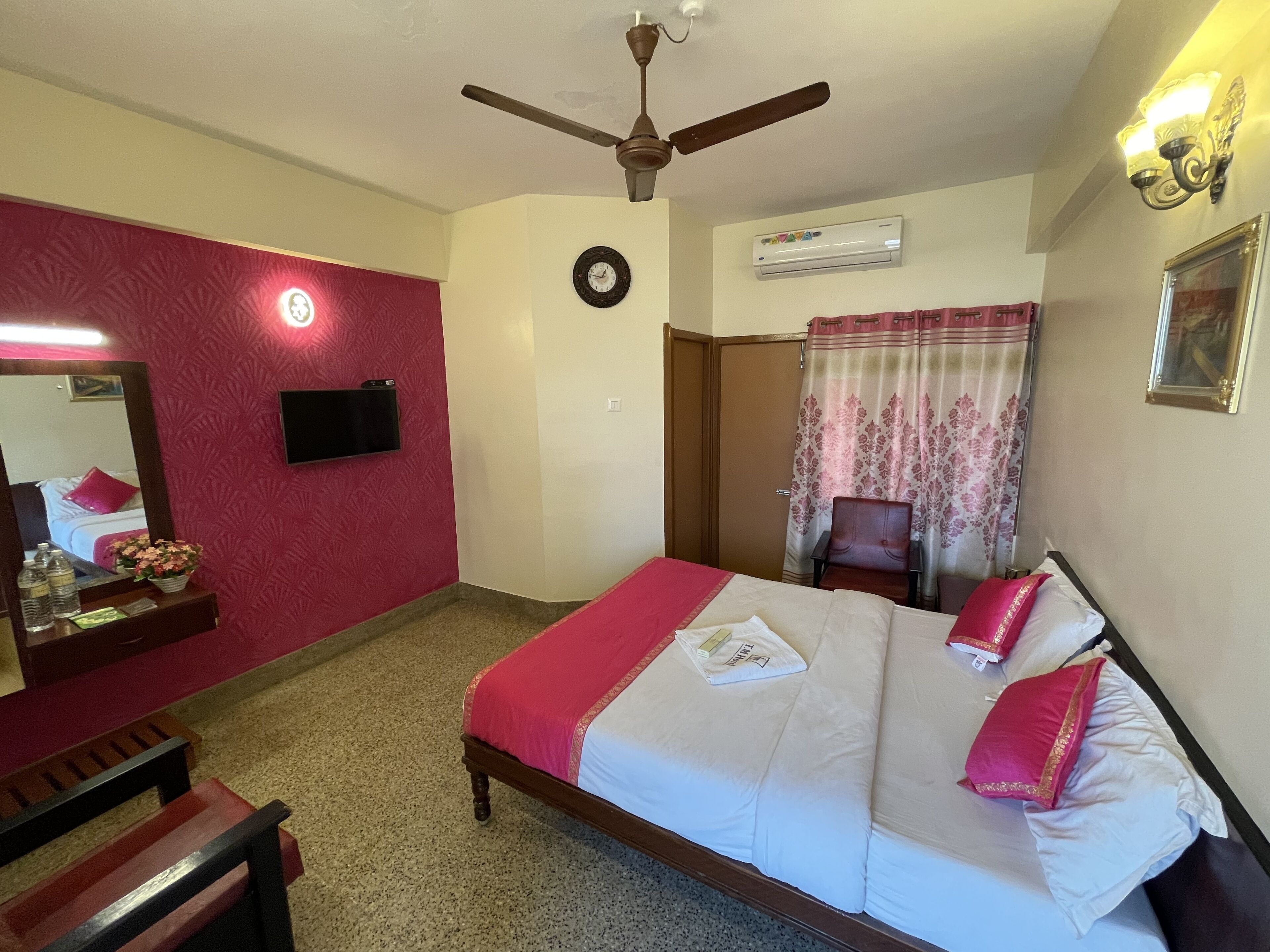 A hotel room with a bed, a ceiling fan, a flat-screen TV, an air conditioning unit, a mirror, a wooden bench, and a floral-patterned curtain.