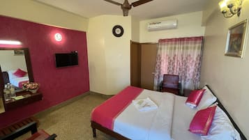 Habitación de hotel con cama, ventilador de techo, televisor de pantalla plana, unidad de aire acondicionado, espejo, banco de madera y cortina con estampado floral.