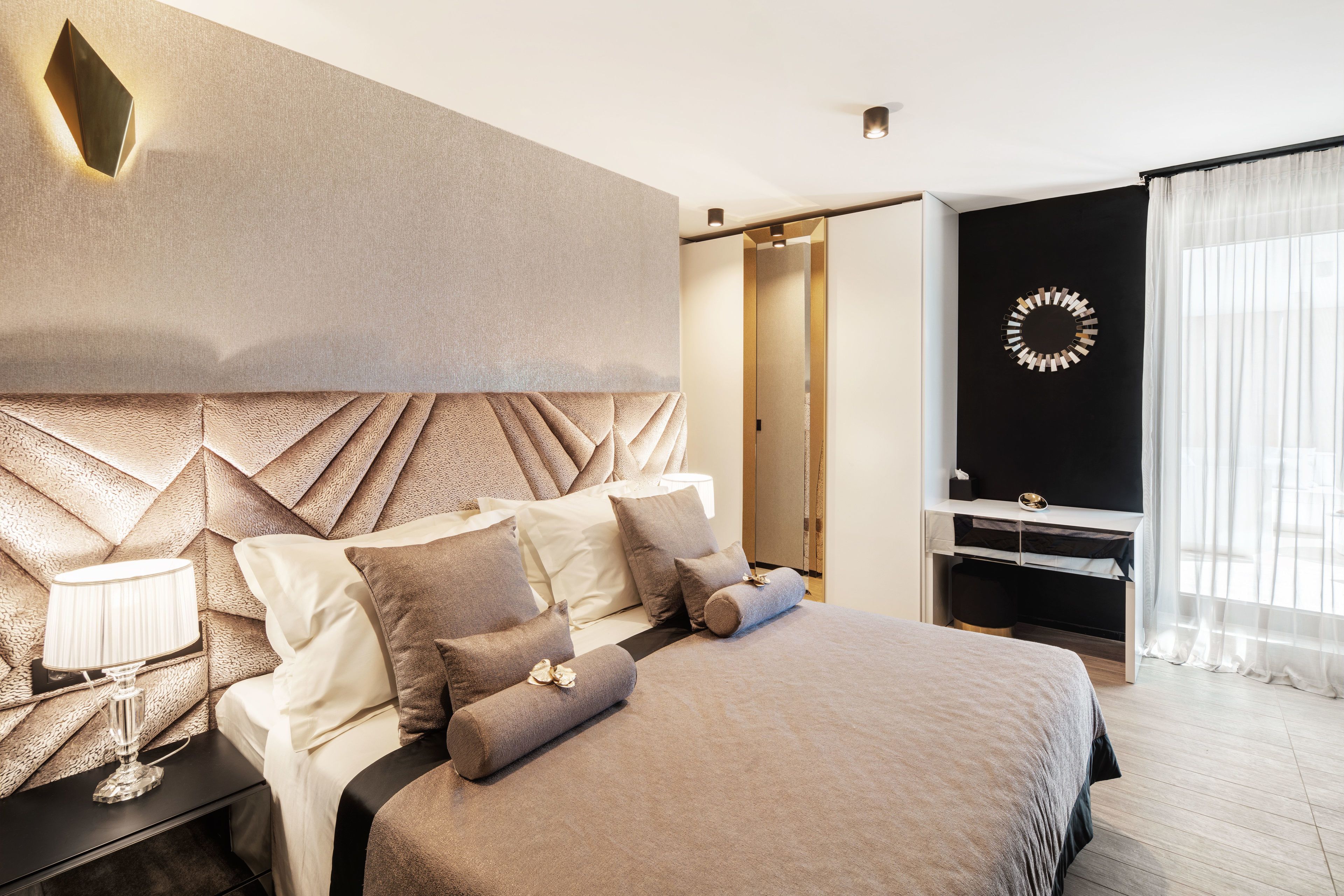 A modern bedroom with a large bed, a nightstand with a lamp, a wall-mounted TV, and a decorative mirror.