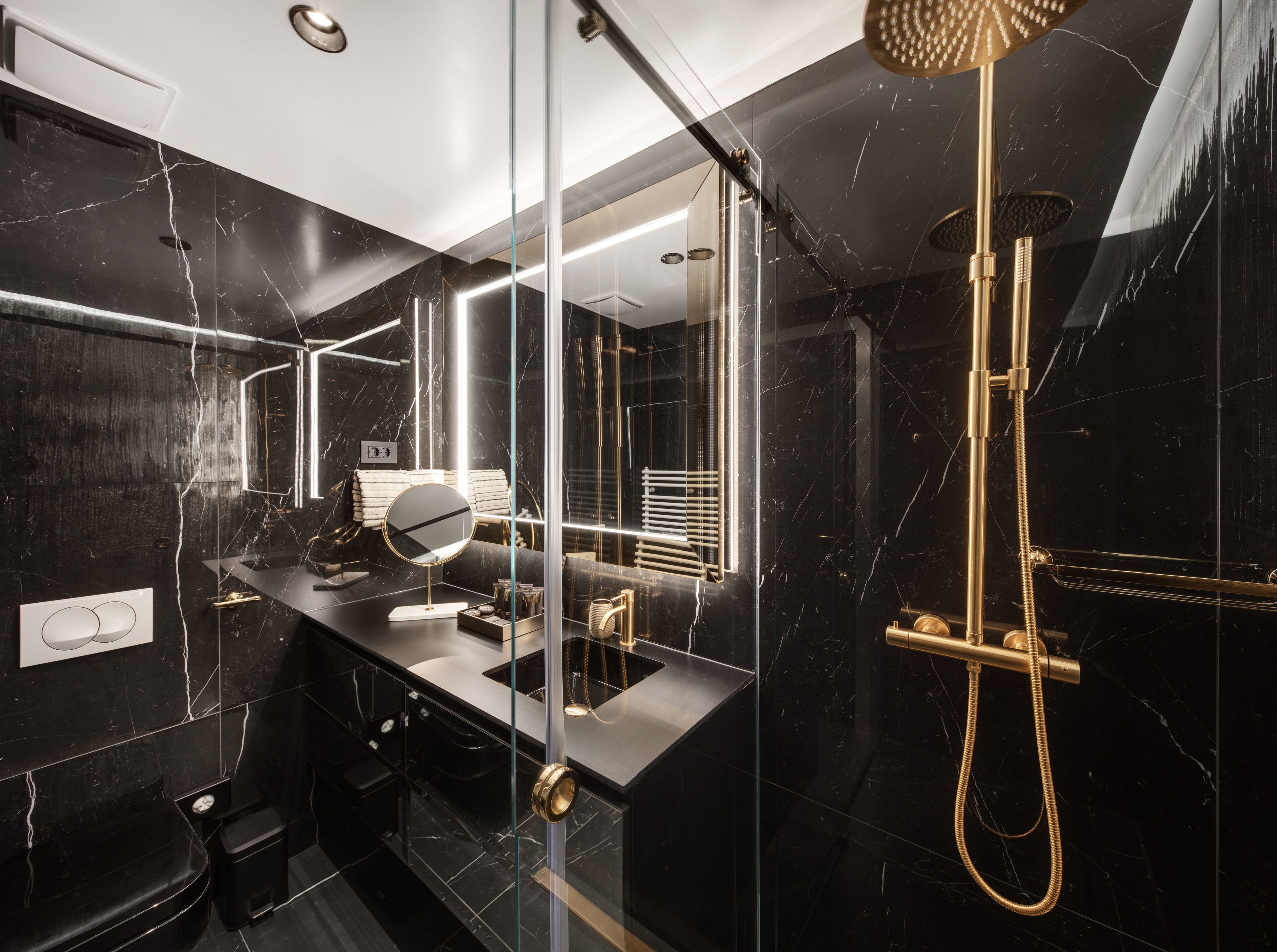 A modern bathroom with a black marble vanity, a round mirror, a gold showerhead, and a glass-enclosed shower.