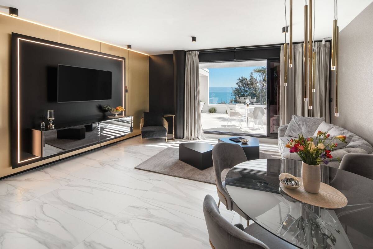 A modern living room with a large flat-screen TV, a glass dining table, and a view of the outdoors.