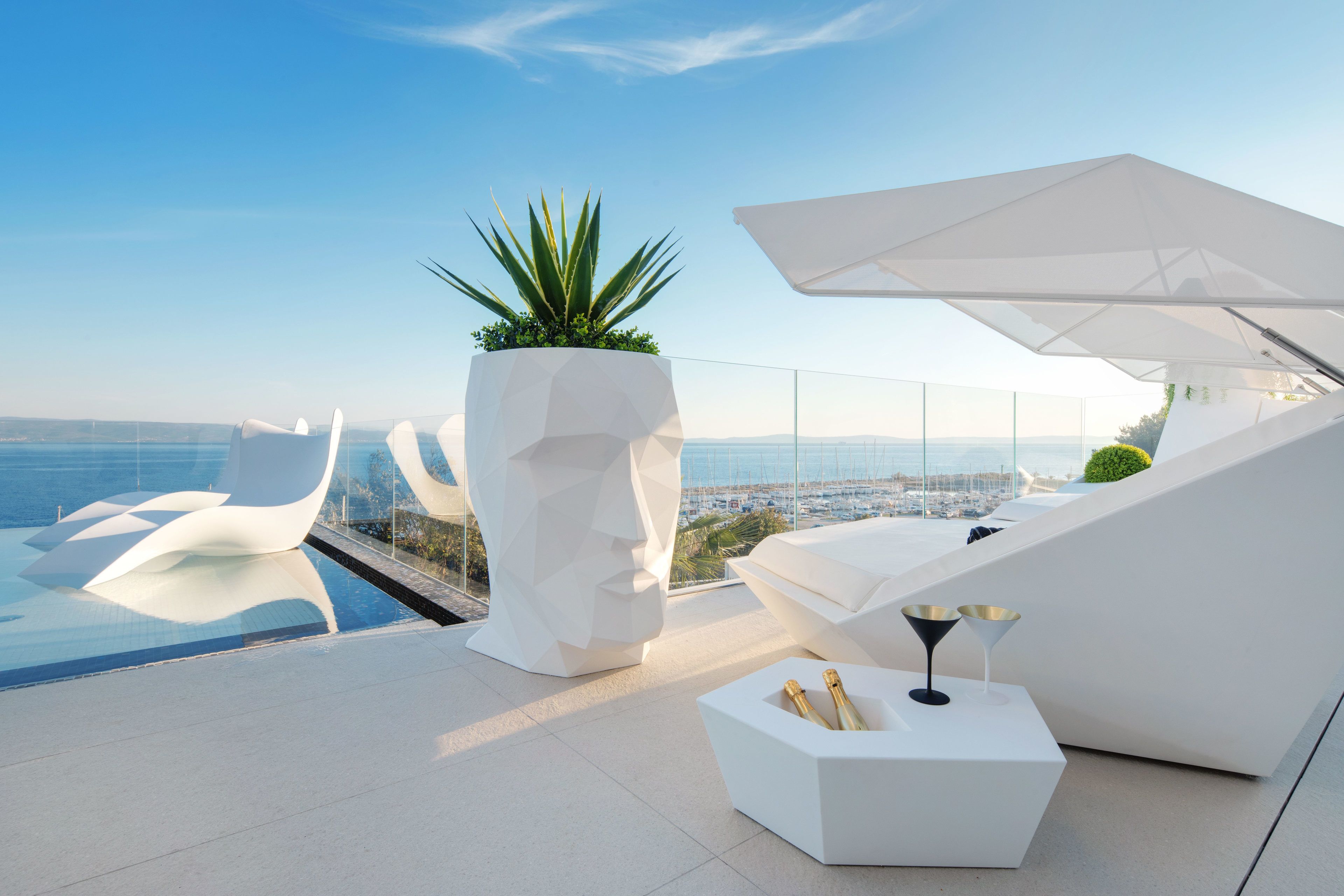 A modern outdoor lounge area with a pool, geometric white furniture, and a view of the sea.