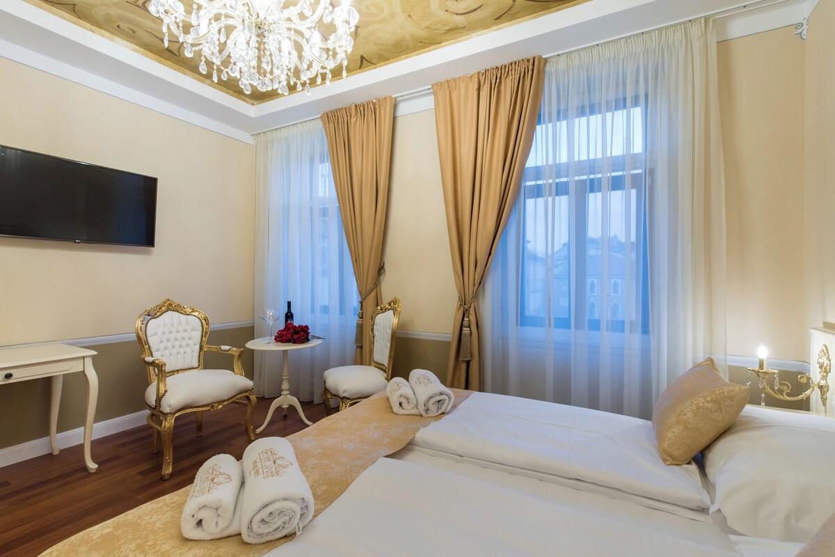 A hotel room with a bed, a TV, a chandelier, a chair, a small table, and a mirror.