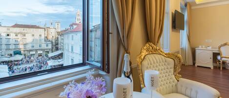 Une chambre d’hôtel luxueuse avec une grande fenêtre donnant sur une place animée, un canapé blanc moelleux, une tête de lit ornée de motifs dorés et une bouteille de champagne Moët.