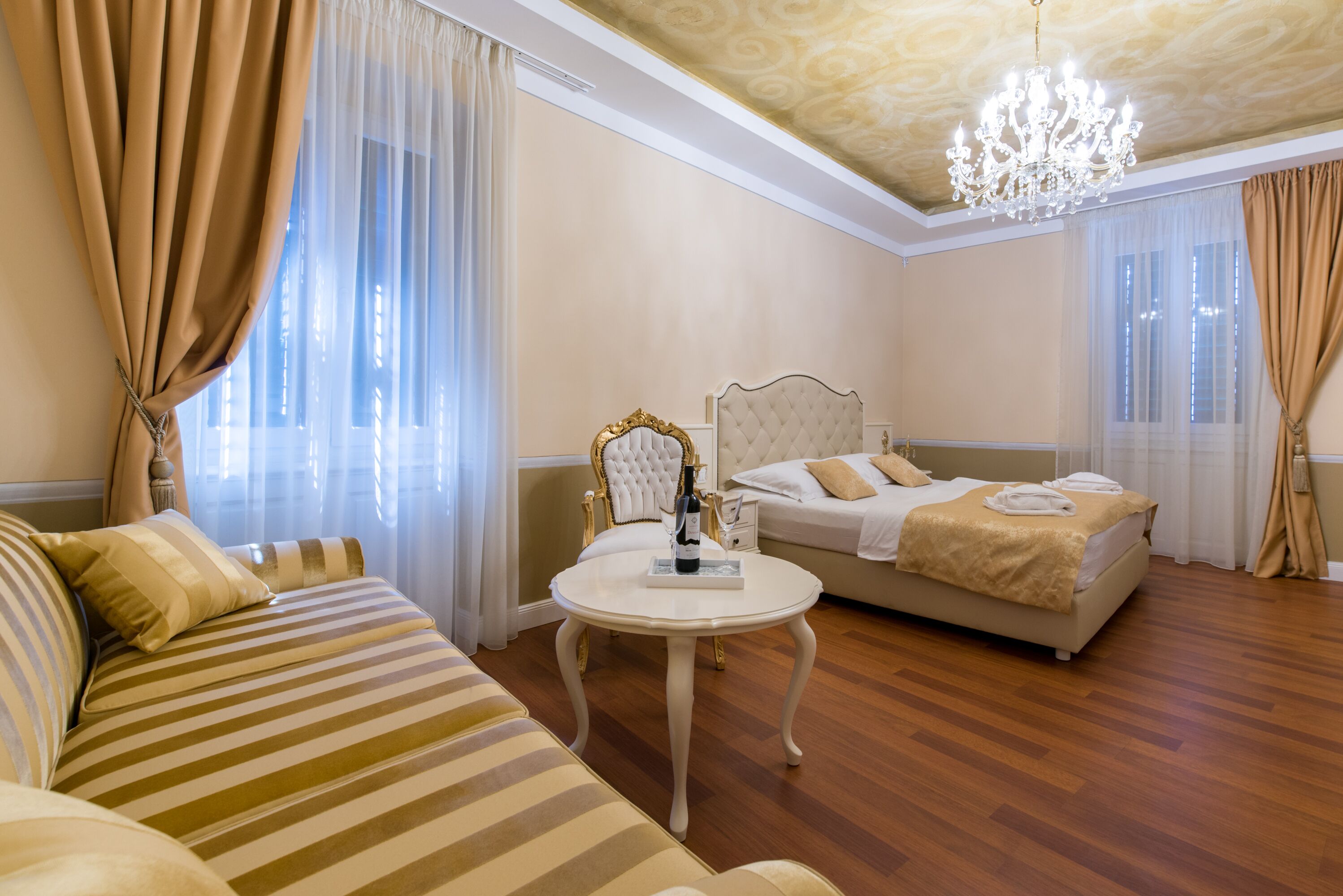 A hotel room with a bed, a sofa, a round table with a bottle, and a chandelier.