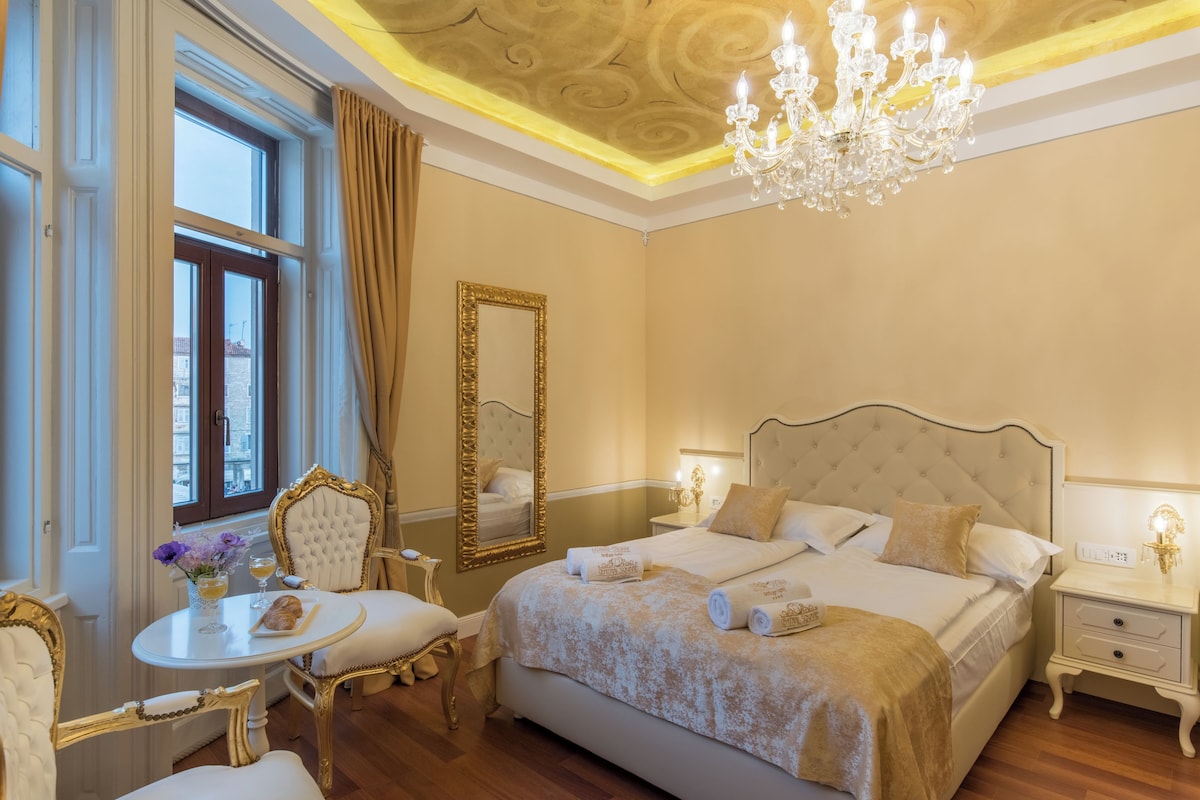 A luxurious bedroom with a large bed, a chandelier, and a dining area with a table and chairs.