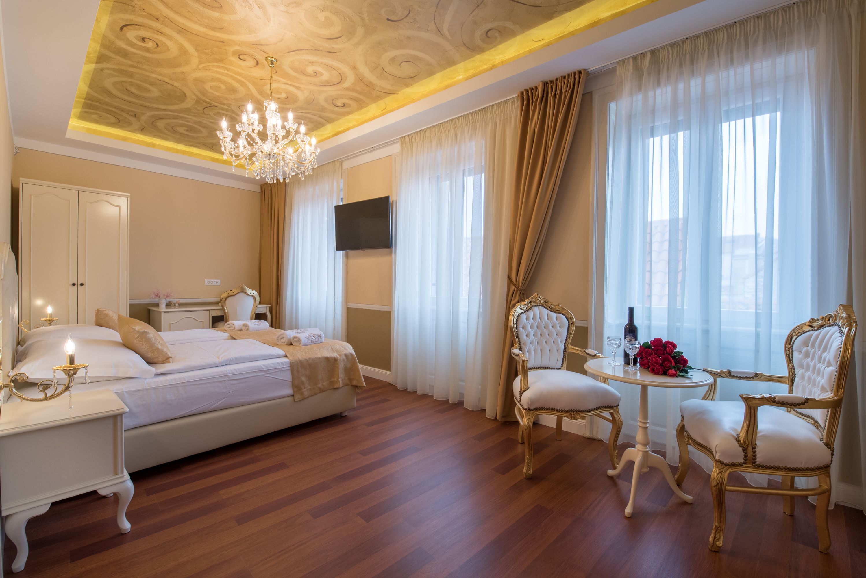 A hotel room with a bed, a chandelier, a TV, a table with chairs, and curtains.