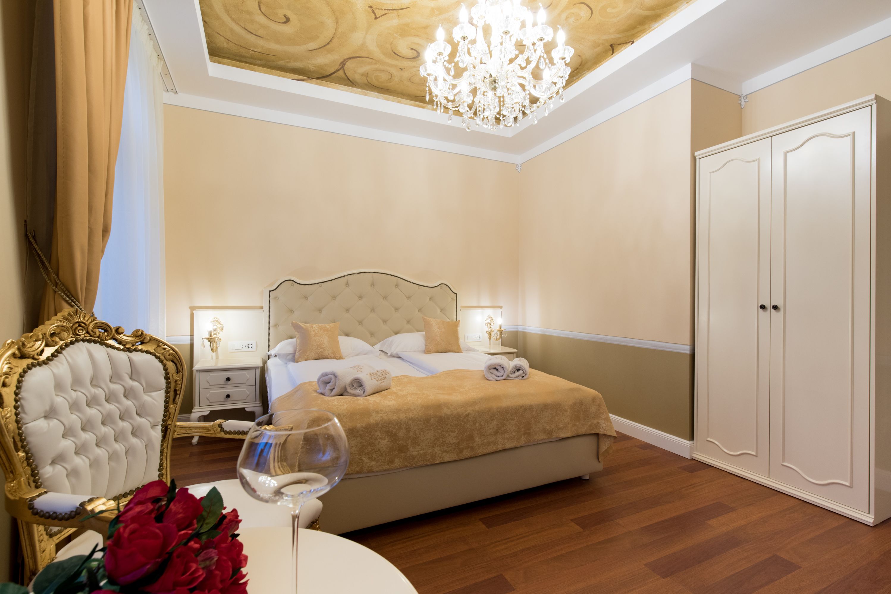A bedroom with a large bed, a chandelier, a wardrobe, and a nightstand with a lamp.
