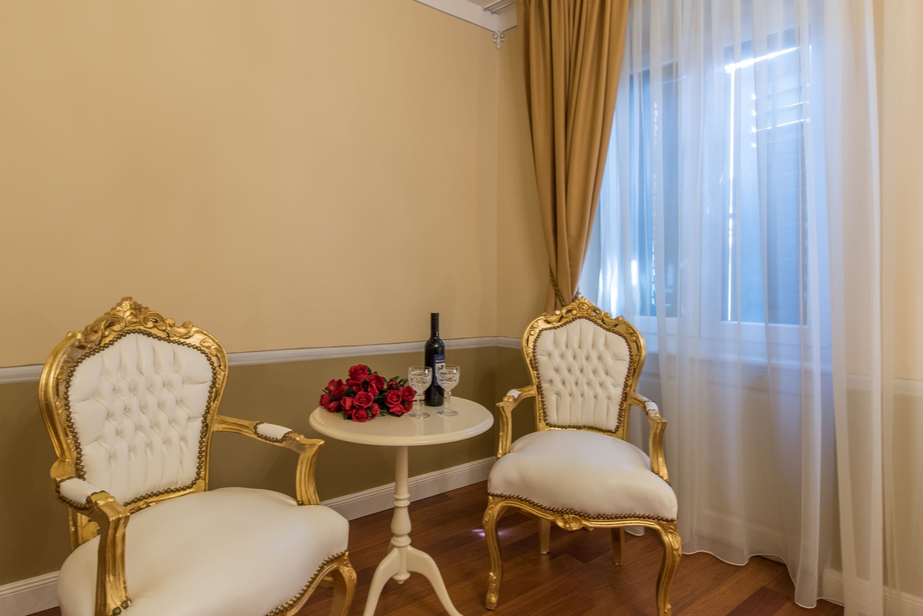 Dos sillones ornamentados con detalles dorados y tapicería blanca, una mesita redonda con una botella y vasos, y un ramo de rosas rojas.