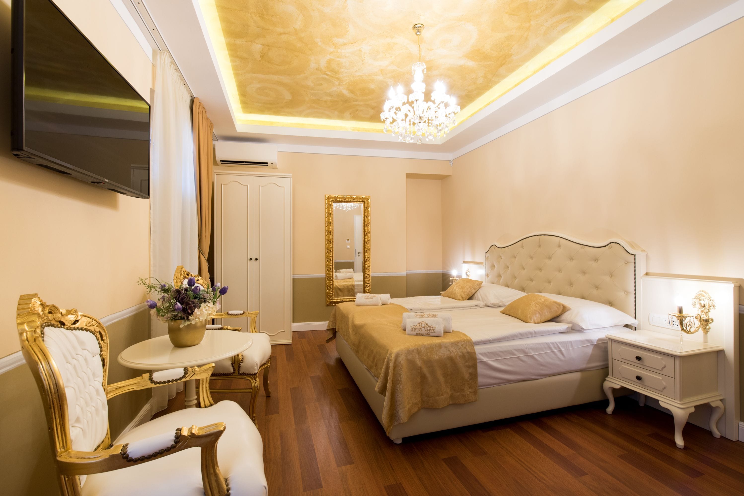 A hotel room with a large bed, a television, a chandelier, and a mirror.