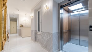 A modern elevator with a metallic finish, a marble wall adjacent to it, and a chandelier with multiple light fixtures. - Central Square Heritage Hotel (Split)