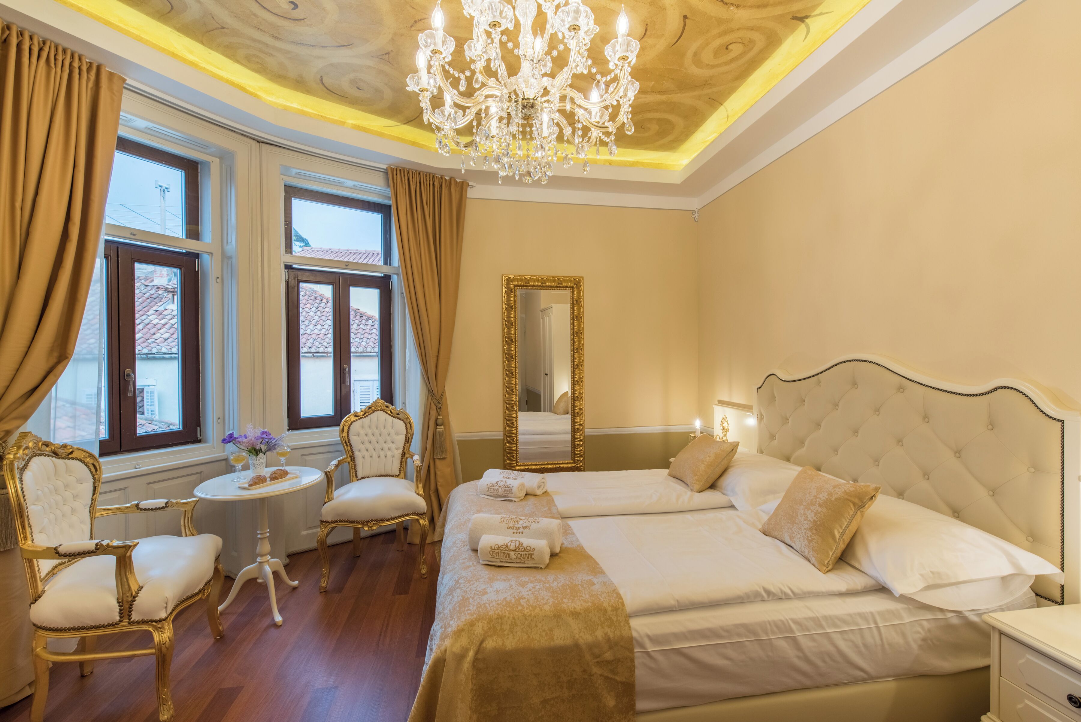 A hotel room with a large bed, two chairs, a round table, a chandelier, and a mirror.