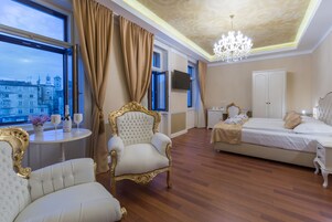 Quarto de hotel luxuoso com uma cama grande, um espaço de estar com poltrona adornada em dourado, uma pequena mesa redonda com um vaso e copos de vinho, e vista para um edifício histórico pela janela.