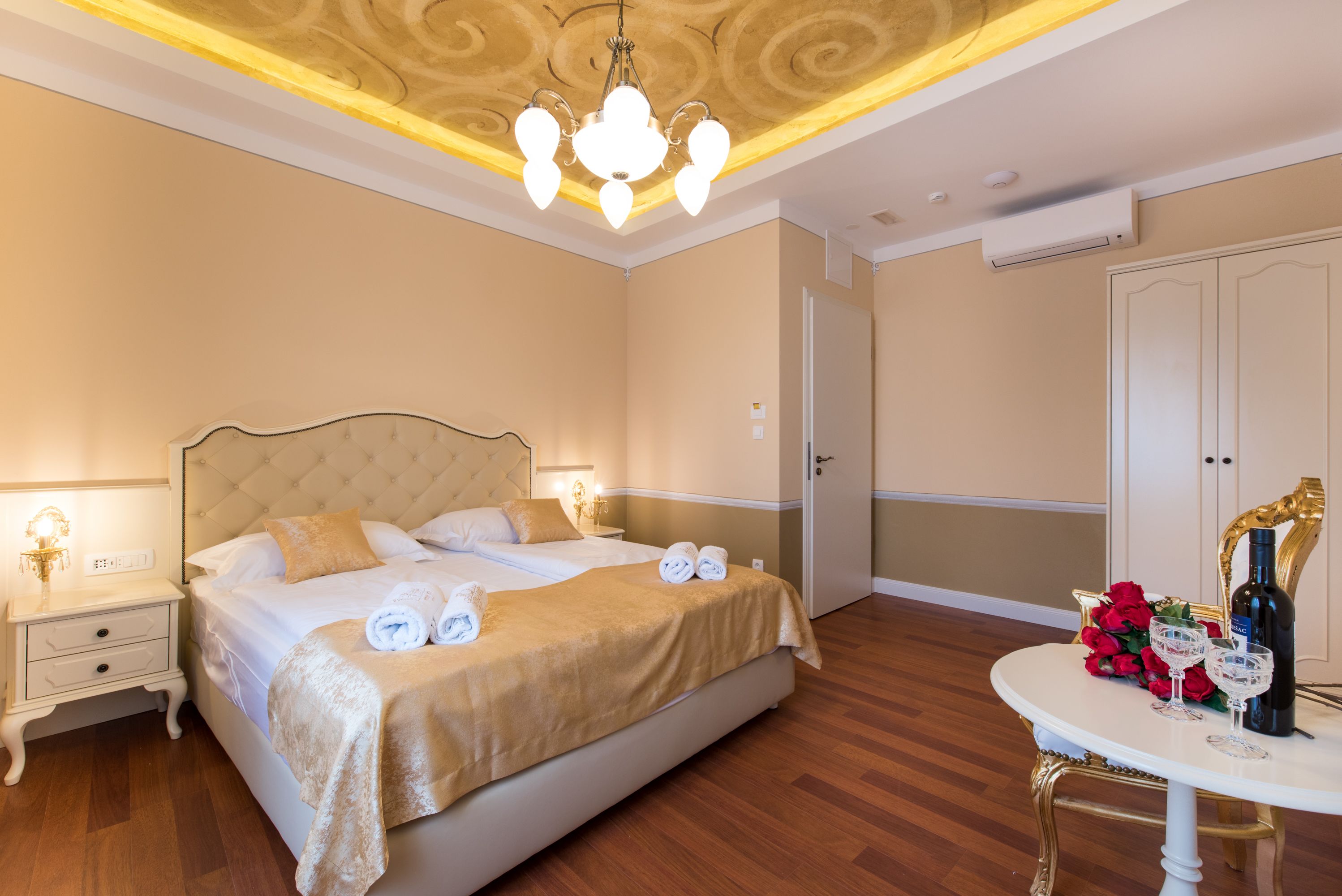 Een hotelkamer met een groot bed, nachtkastjes, een kroonluchter en een tafel met een fles en glazen.