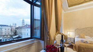 A hotel room with a large window offering a city view, a bed with a gold headboard, and a small table with a bouquet of red roses and a wine glass. - Central Square Heritage Hotel (Split)