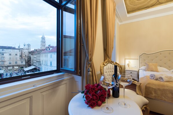 A hotel room with a large window, a bed with a golden headboard, a bedside table with a wine glass and a bottle, and a bouquet of red roses.