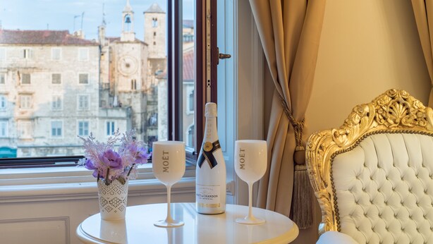 A table with a bottle of Moët champagne and two glasses, a vase with flowers, and a view of historic buildings through a window. - Central Square Heritage Hotel (Split)