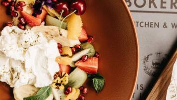 Un plato de ensalada de frutas con yogur y semillas de granada, un vaso de jugo de naranja y una cuchara de madera sobre una mesa con un periódico.
