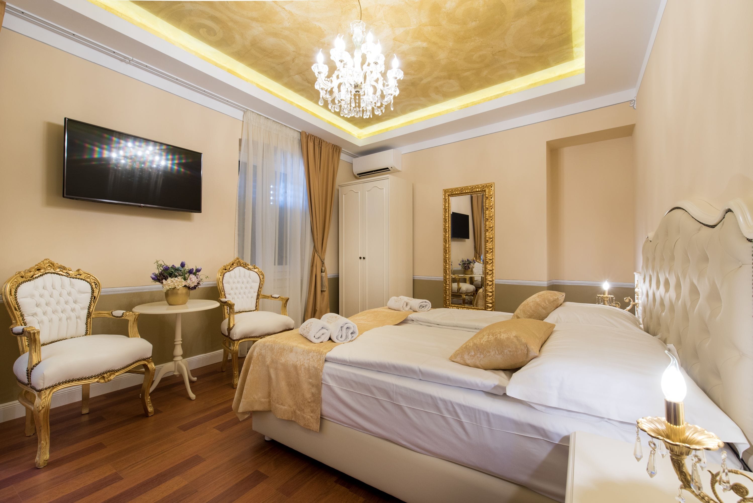 A spacious bedroom with a large bed, two ornate chairs, a small round table with a vase, a television, and a mirror.