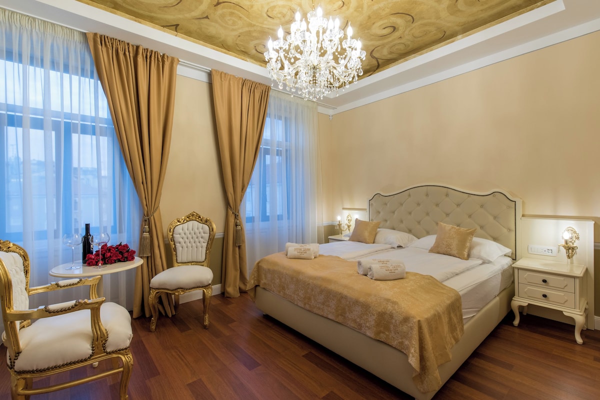 A luxurious bedroom with a large bed, a chandelier, and a view of the city through large windows.