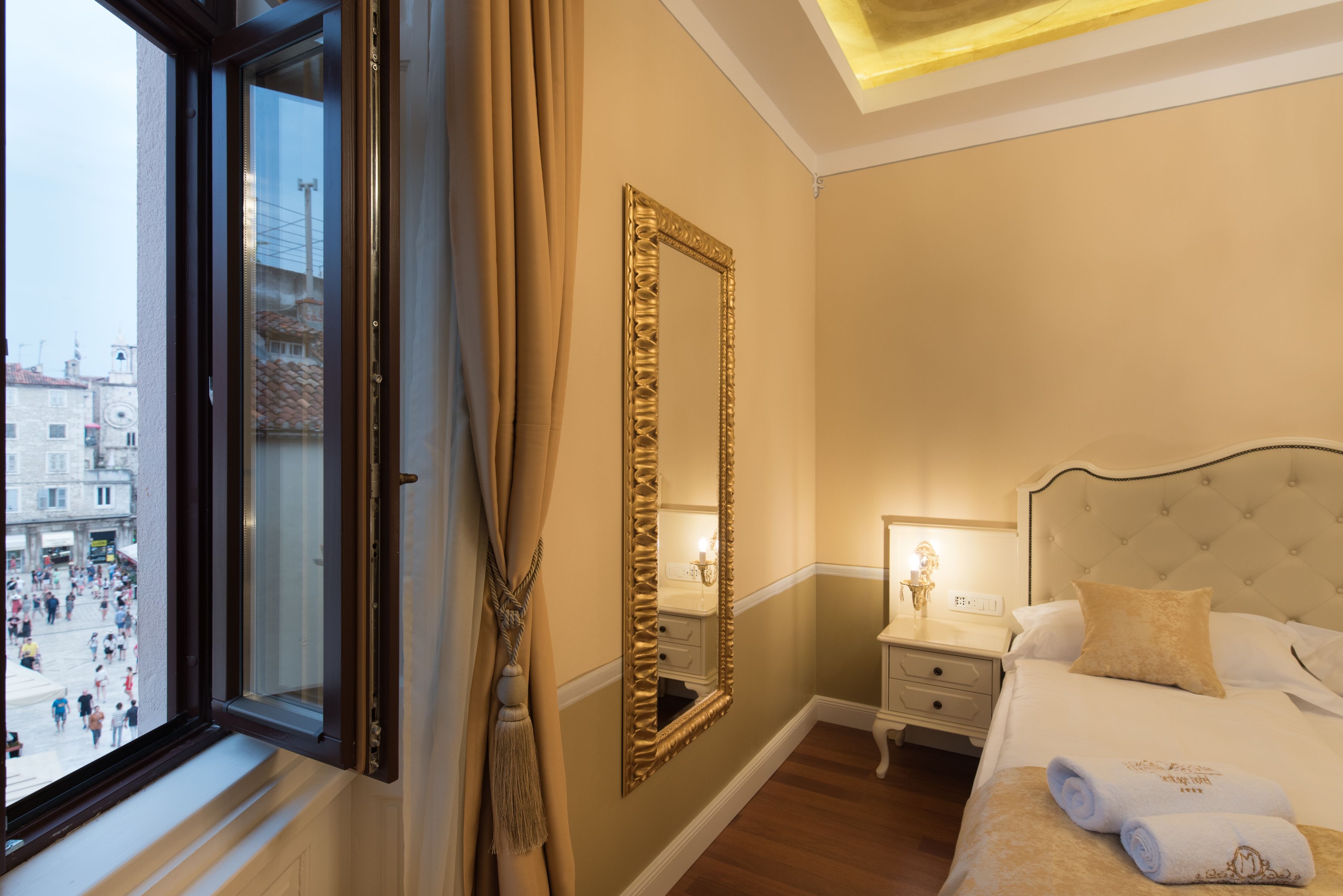Habitación de hotel con un espejo grande, una cama con ropa blanca, una mesita de noche y vistas a una calle con movimiento a través de una ventana.