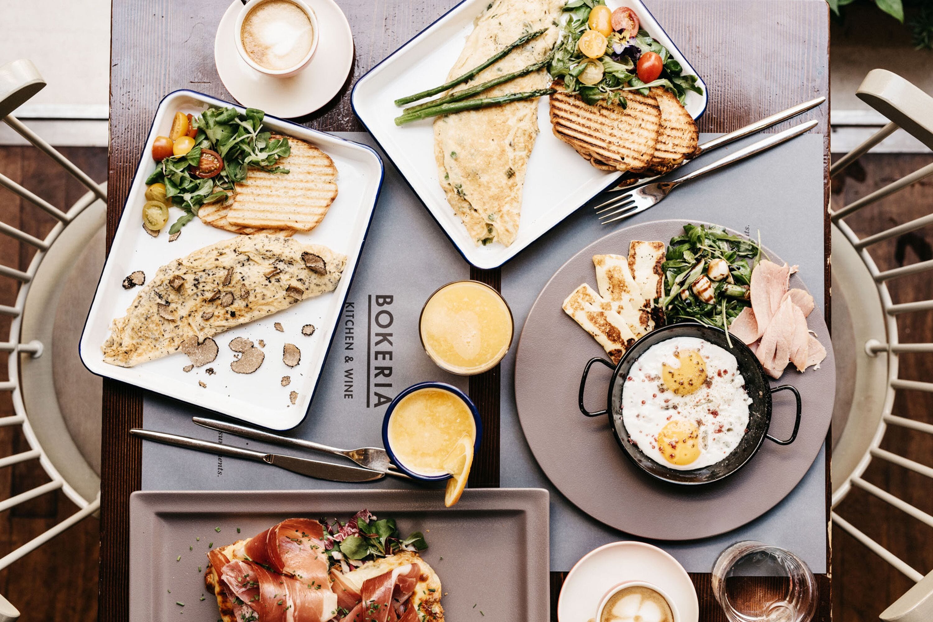 A dining table with various dishes including grilled fish, eggs, and salads, along with cups of coffee and a menu titled "BOKERIA".