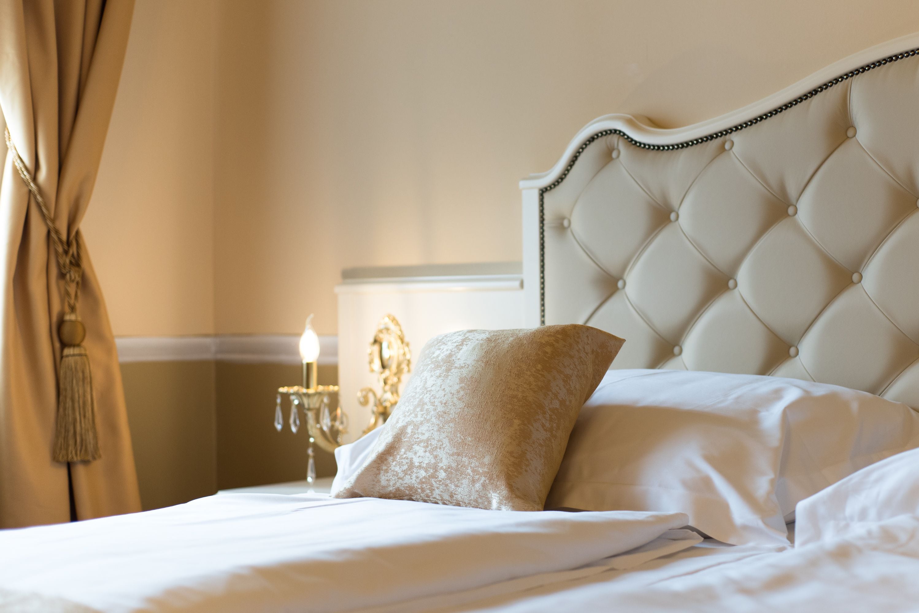 A neatly made bed with a tufted headboard, a decorative pillow, and a chandelier in the background.