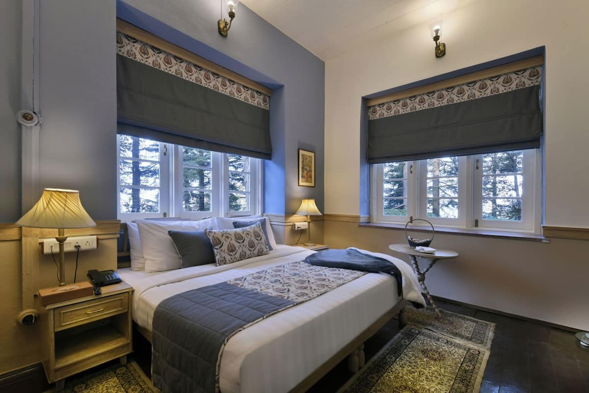 Un dormitorio con cama, lámparas de noche, mesita de noche, una pequeña mesa y una ventana con vistas a árboles.