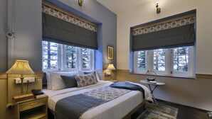 Un dormitorio con dos camas, una mesita de noche con lámpara, una pequeña mesa con una cesta y una ventana con vistas a árboles.