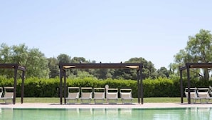 Área de piscina com espreguiçadeiras sob pérgulas, cercada por vegetação.