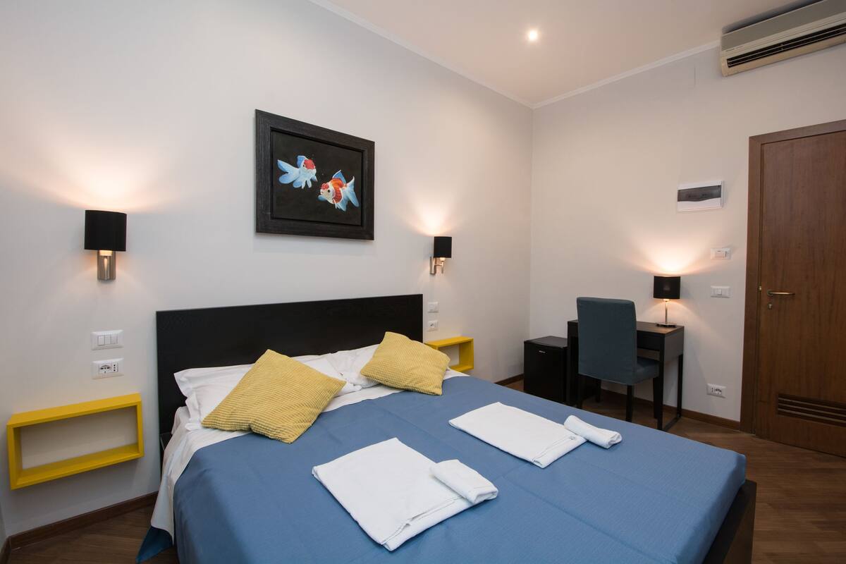 Ein ordentlich bezogenes Bett mit blau-weißer Bettwäsche, zwei Kissen und gefalteten Handtüchern. Ein Schreibtisch mit Stuhl, ein gerahmtes Bild an der Wand und eine Tür auf der rechten Seite sind ebenfalls zu sehen.
