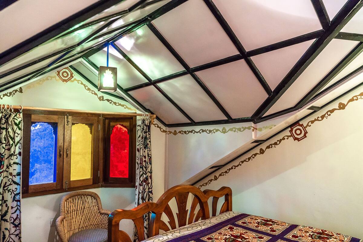 A room with a sloped ceiling, a wooden chair, a table with a patterned tablecloth, and a window with stained glass panes.