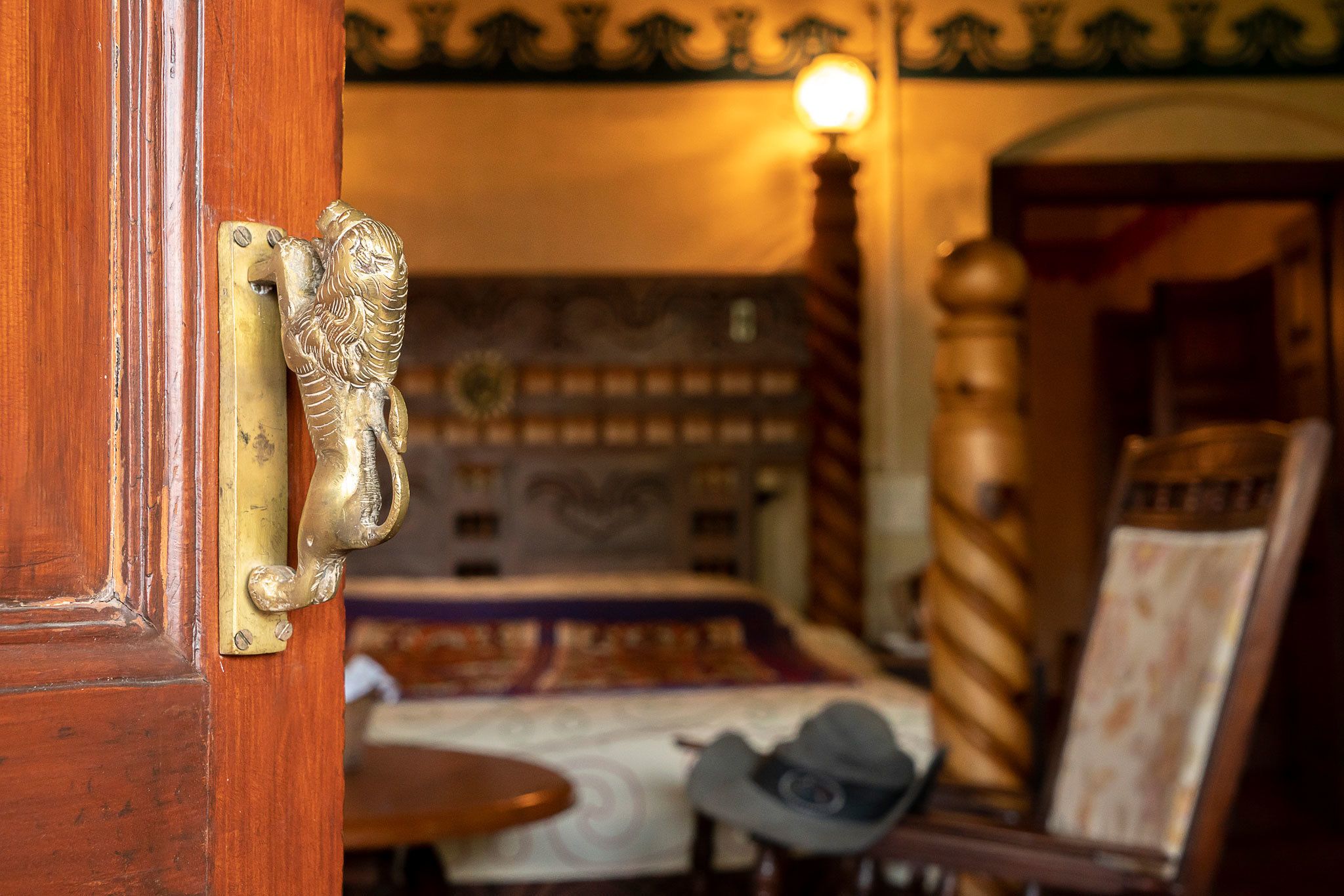 A wooden door with a lion-shaped handle, a patterned wall, a bed with a patterned bedspread, a round table, and a chair with a hat.