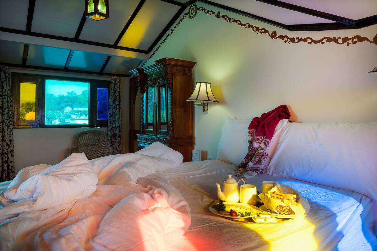 A neatly made bed with a tray of breakfast items, a lamp, and a window with a view of a building.