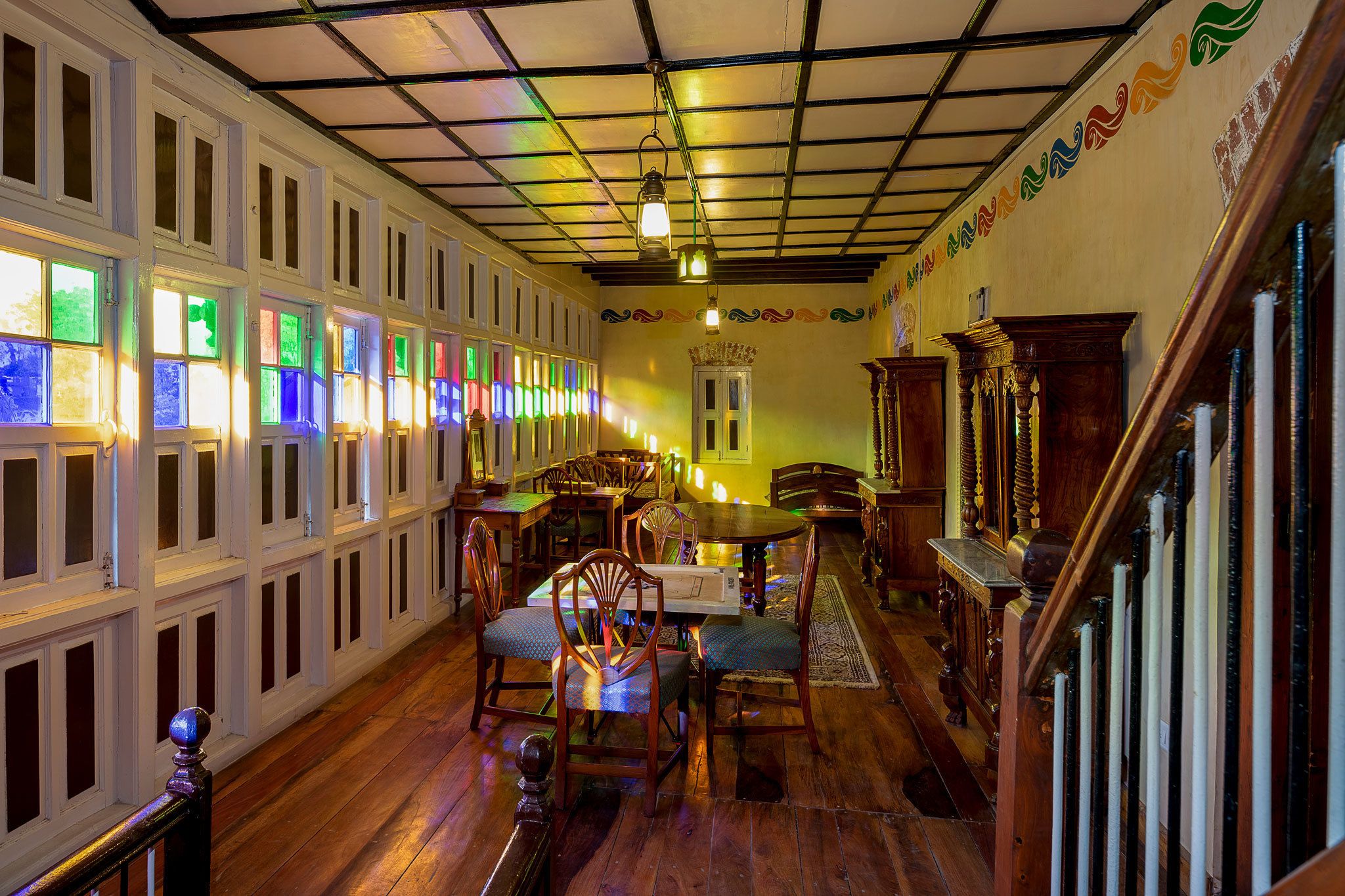 A room with a wooden floor, white paneled walls, a dining table with chairs, and a staircase with a railing.