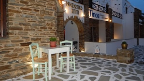 Un edificio de piedra con un letrero que dice "NHSTIKO EMITTI" y "Tinos Suites & Apartments". Hay zonas de estar al aire libre con sillas y mesas, y plantas en macetas.