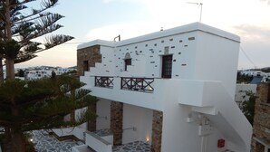 Un edificio moderno de color blanco con base de piedra, un balcón y un patio con pavimento de piedra y plantas en macetas.