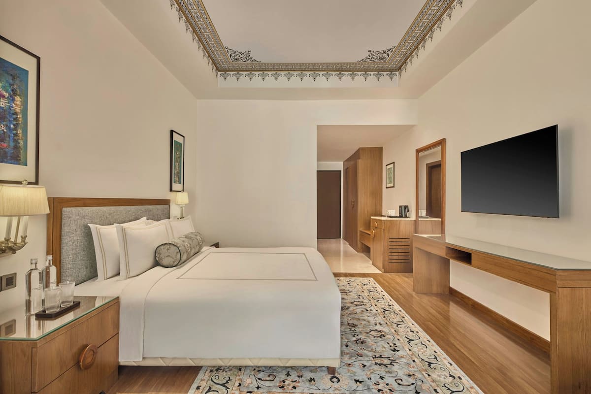 A hotel room with a large bed, a flat-screen TV, a wooden wardrobe, and a decorative ceiling.