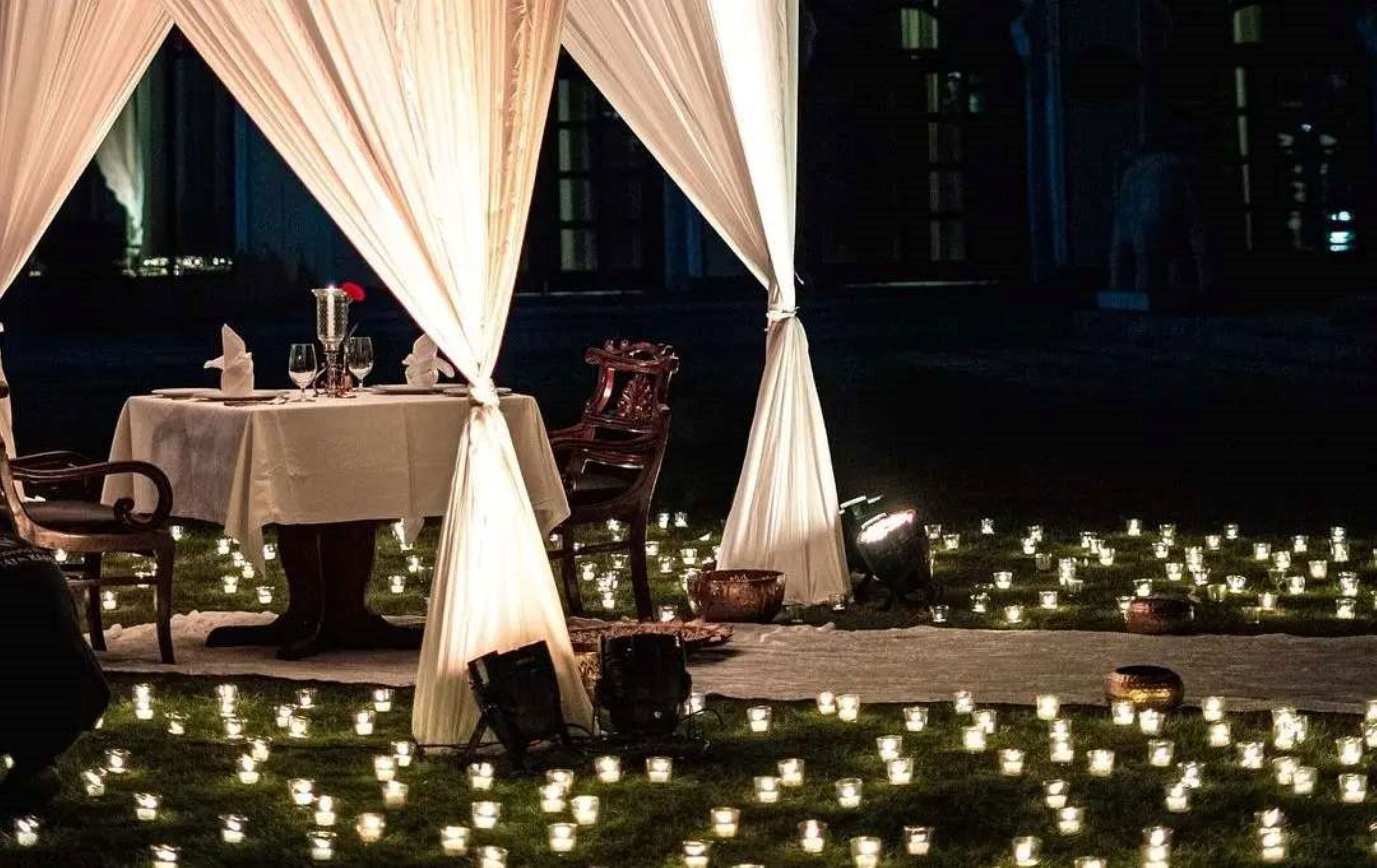 Un área de comedor al aire libre con una mesa preparada para dos personas, rodeada de cortinas blancas y velas sobre el césped.