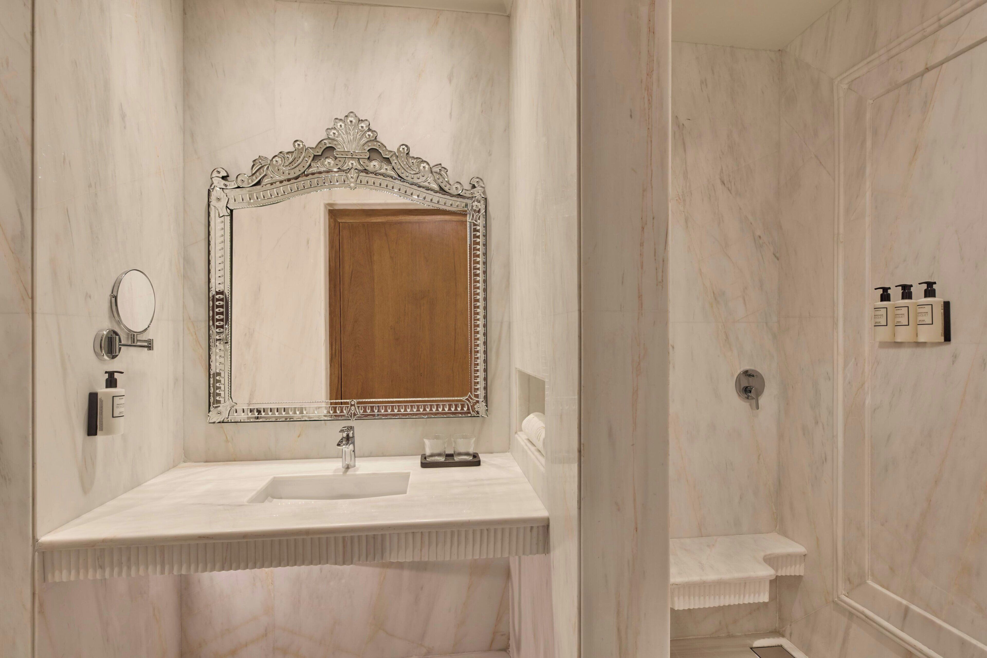 Un bagno con un mobile lavabo in marmo, uno specchio grande e decorato, un armadietto in legno, una zona doccia e un porta asciugamani.