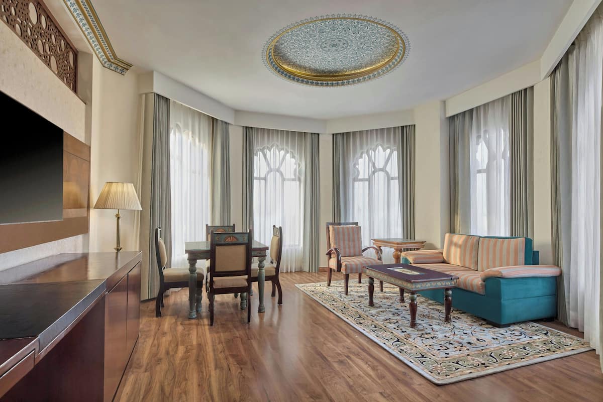 A hotel room with a wooden floor, a sofa, a dining table with chairs, a TV, and a decorative ceiling.