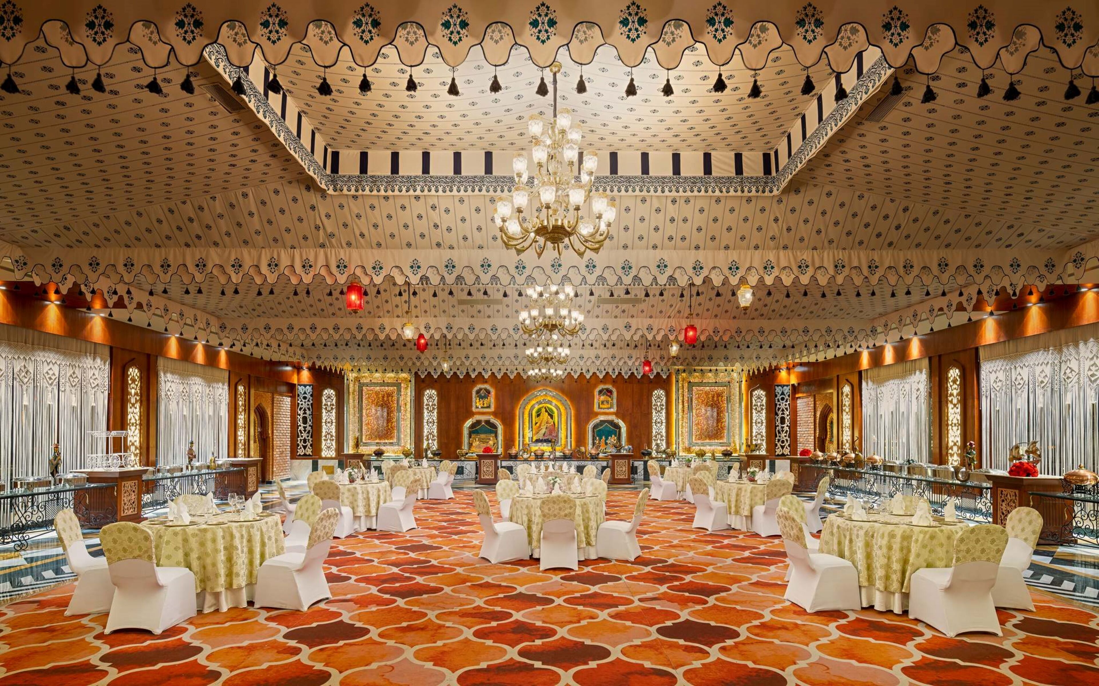 A banquet hall with ornate ceiling patterns, chandeliers, and a patterned floor. Tables are set with white tablecloths and chairs.