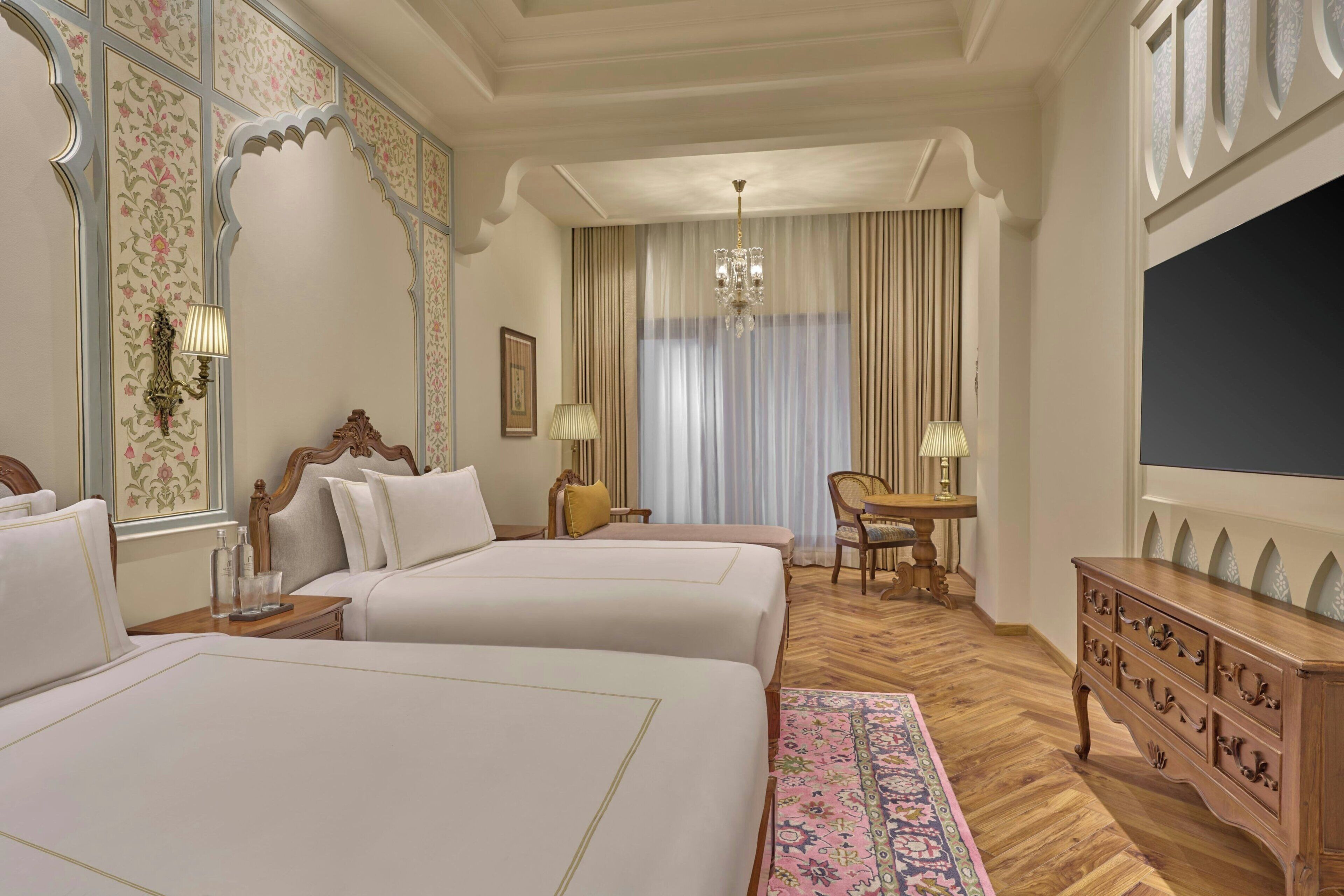 A hotel room with two beds, a wooden dresser, a desk with a chair, a chandelier, and a large window with curtains.