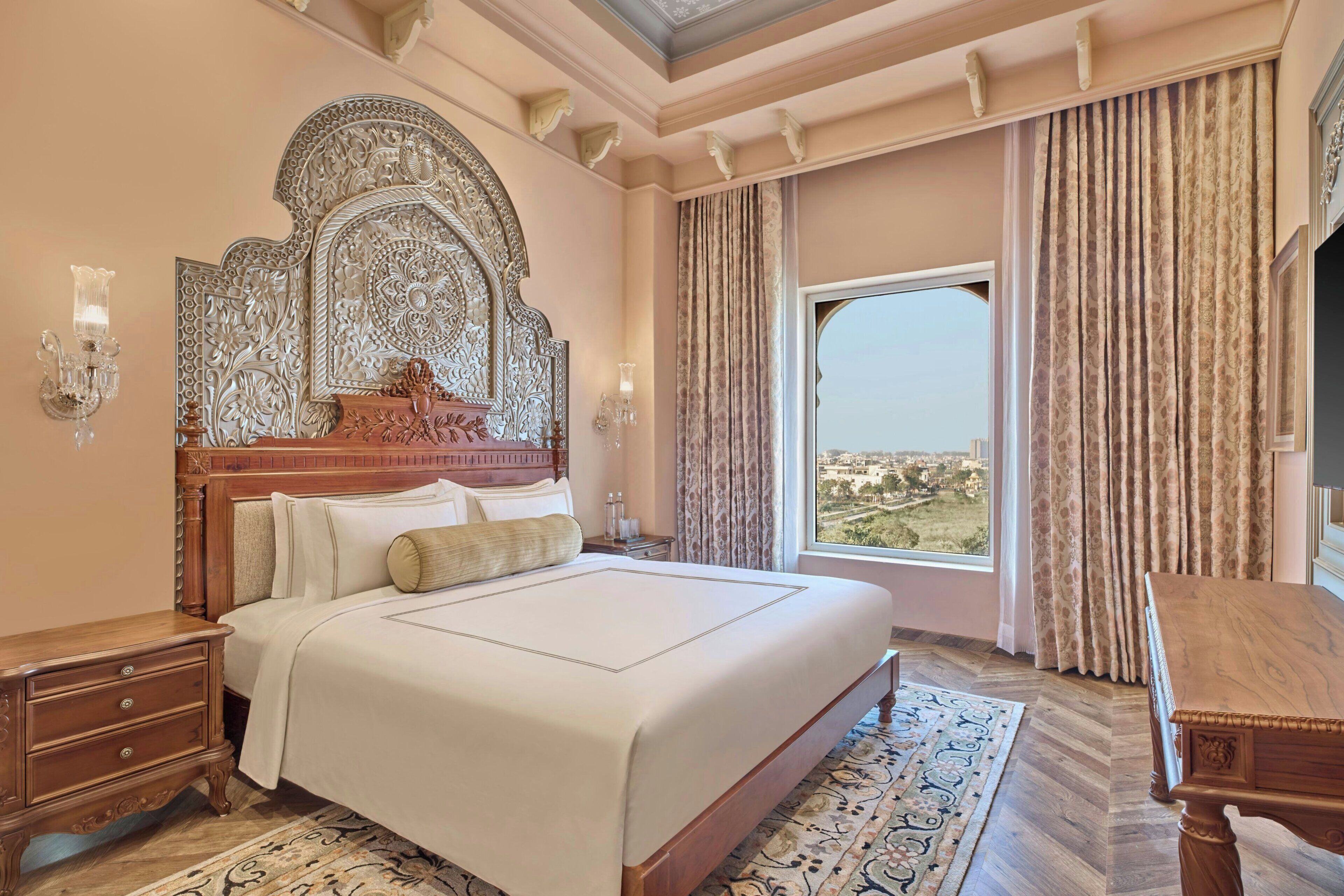 A hotel room with a large bed, ornate headboard, a nightstand, a wooden dresser, a patterned rug, and a large window with curtains.