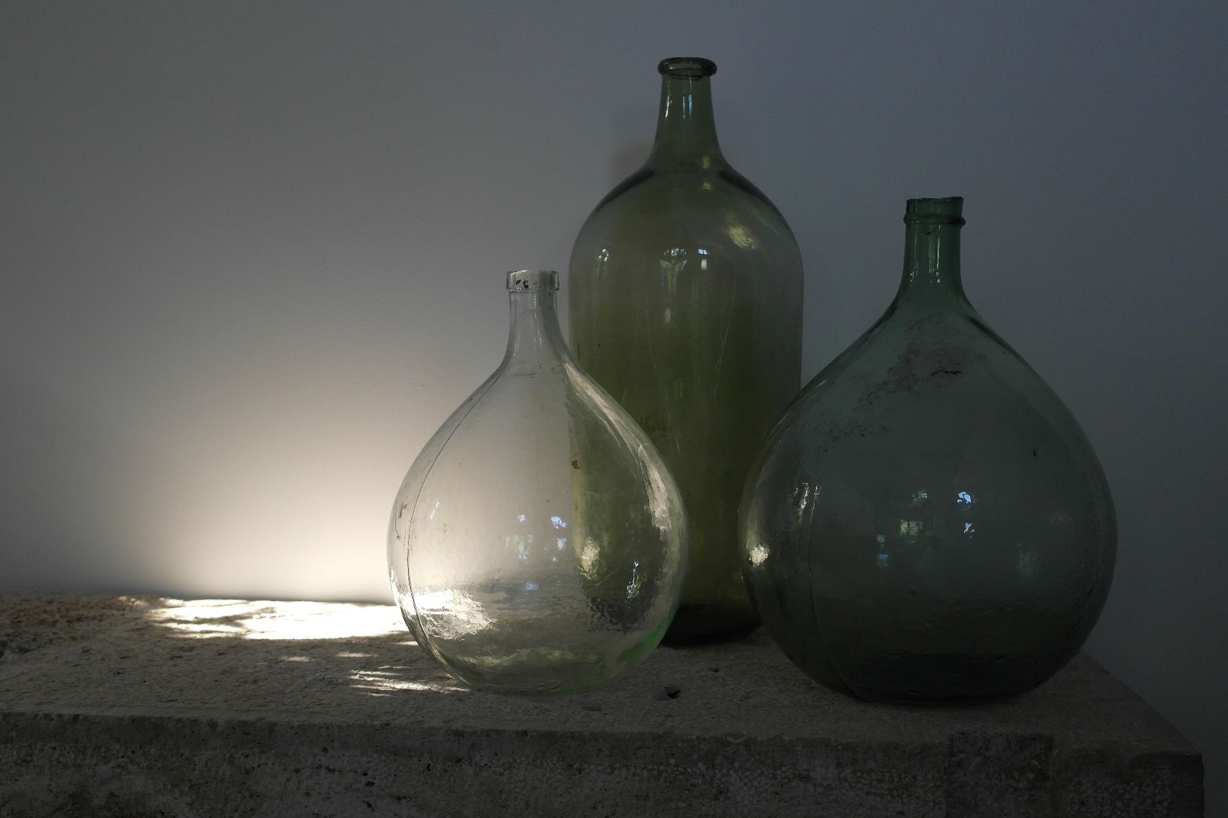 Three glass bottles of varying sizes on a concrete ledge against a plain wall.