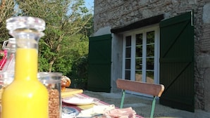 A stone building with a window and green shutters, a table with fruits and a bottle, and a bench in the foreground. - Les chambres de Filou (Montesquieu)